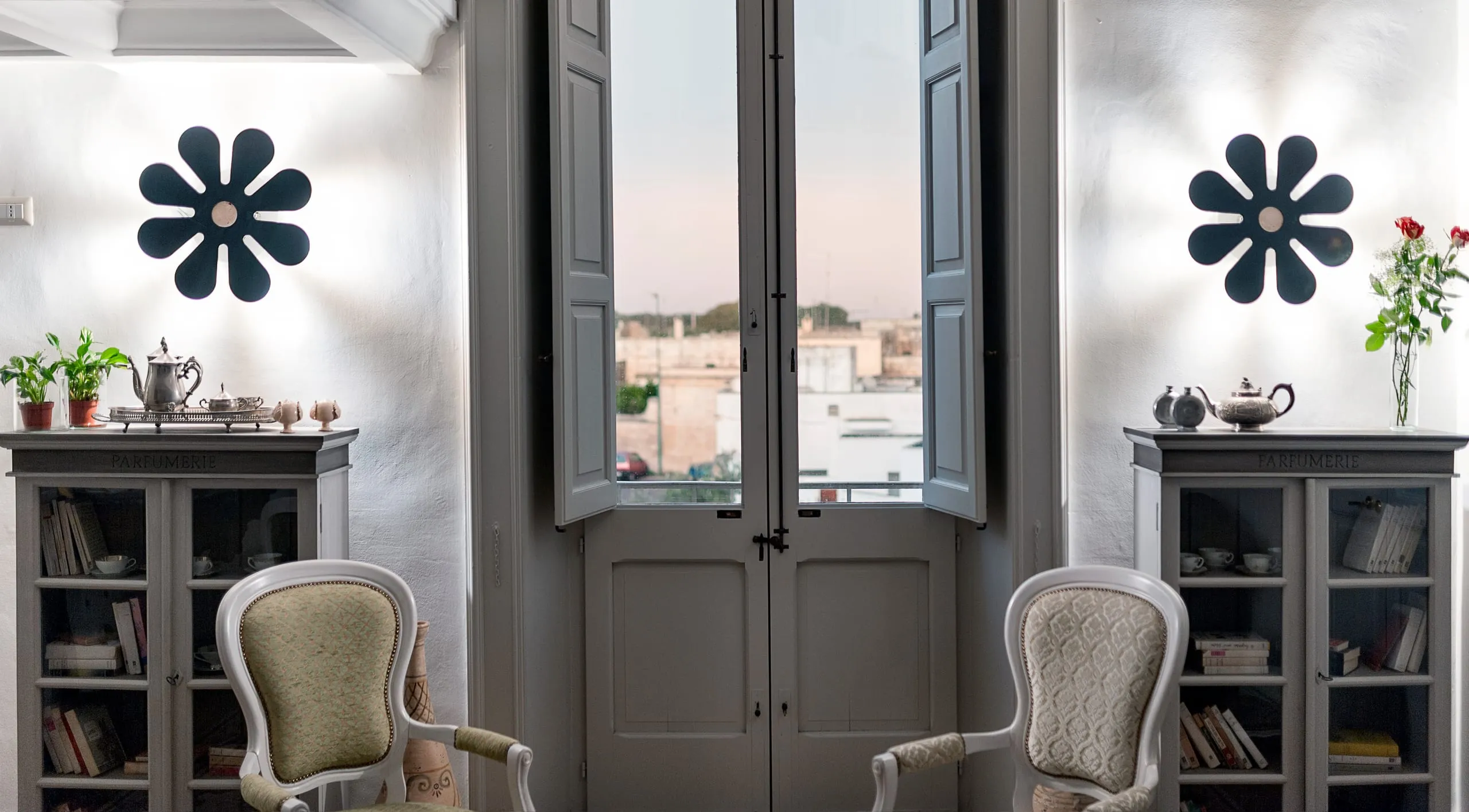 La sala delle finestre di Palazzo Mandurino al tramonto: la portafinestra aperta sui tetti di Zollino fra due vetrine di libri, le due appliques a fiore accese sulle pareti bianche e due poltroncine imbottite.