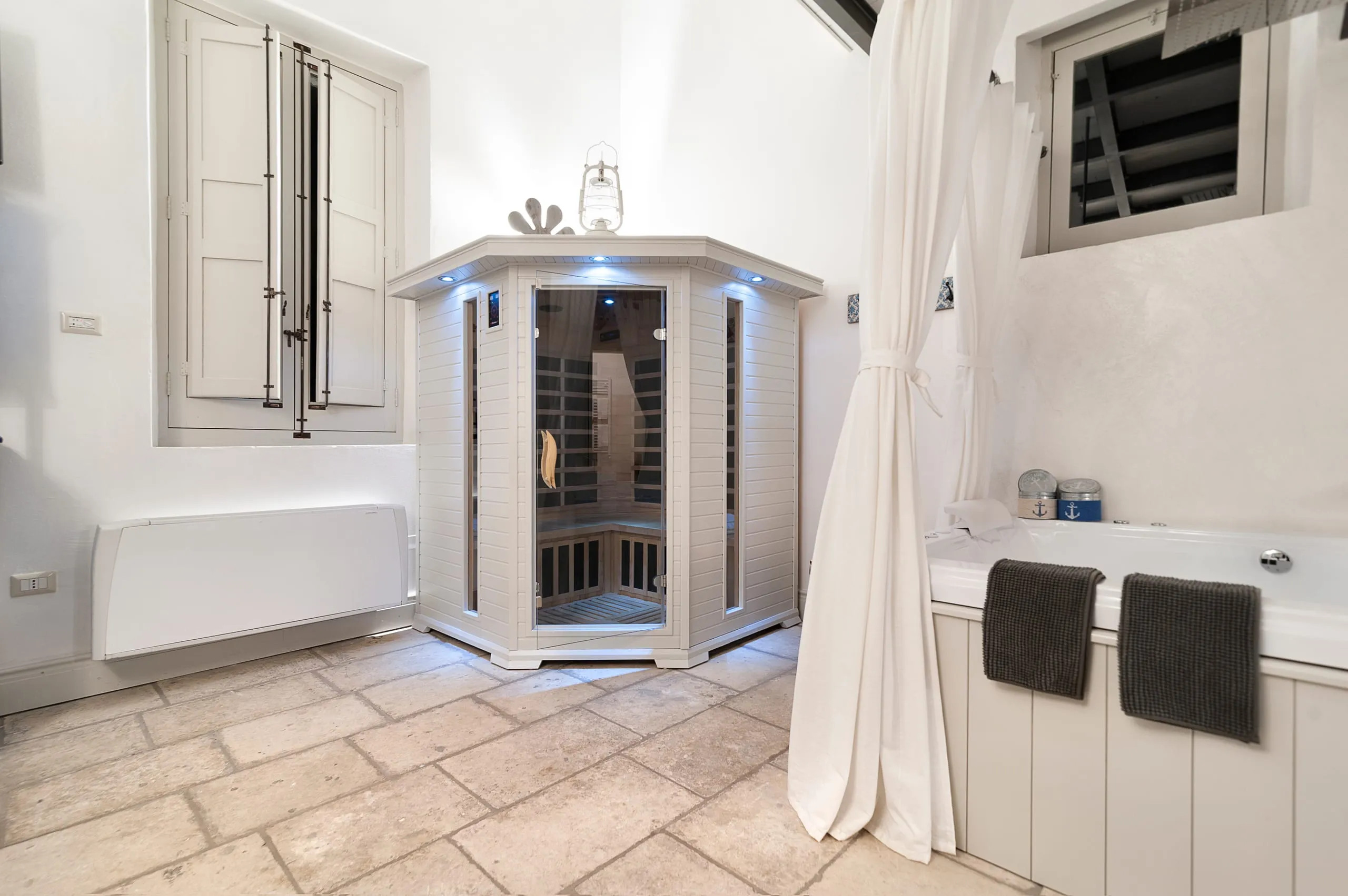 La cabina della sauna a infrarossi della suite Ascilò, accesa, accanto alla vasca idromassaggio e alla finestra con le persiane chiare.