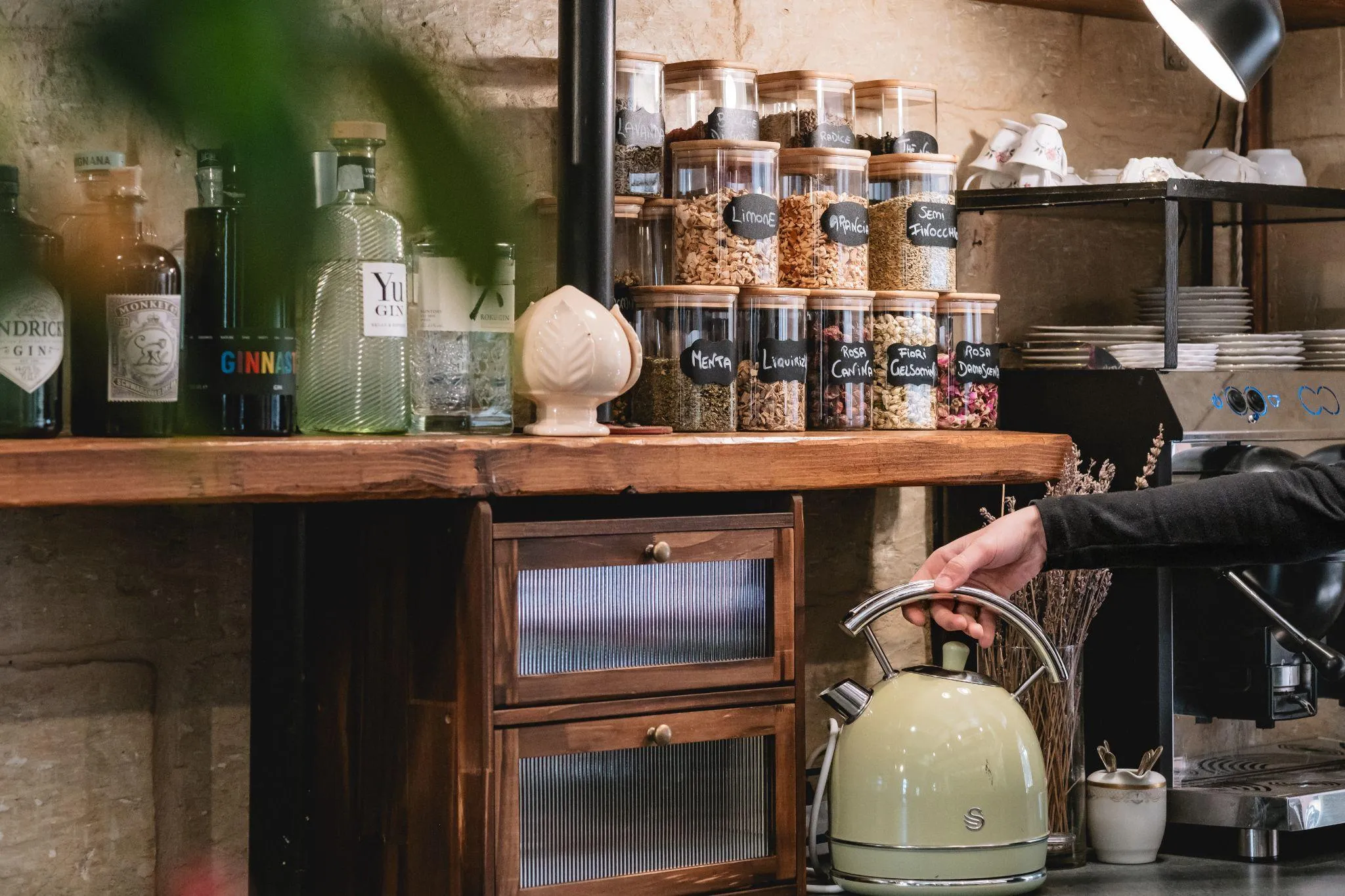 Il bancone del lounge bar: i barattoli di infusi con le etichette scritte a mano — menta, liquirizia, rosa canina — sopra il piano di legno massello, accanto alla macchina del caffè.