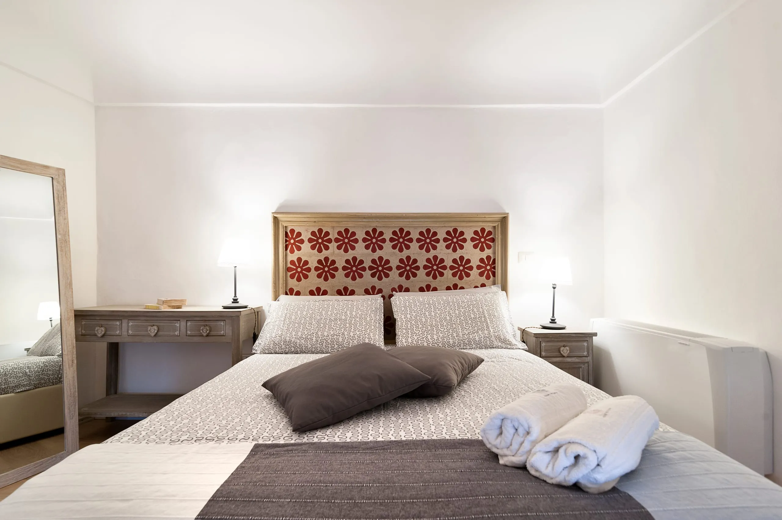 La camera della suite Casa Grika: il letto con la testiera dorata a fiori rossi ricamati, i due comodini di legno chiaro con le lampade accese, gli asciugamani arrotolati sul copriletto e uno specchio alto appoggiato alla parete.