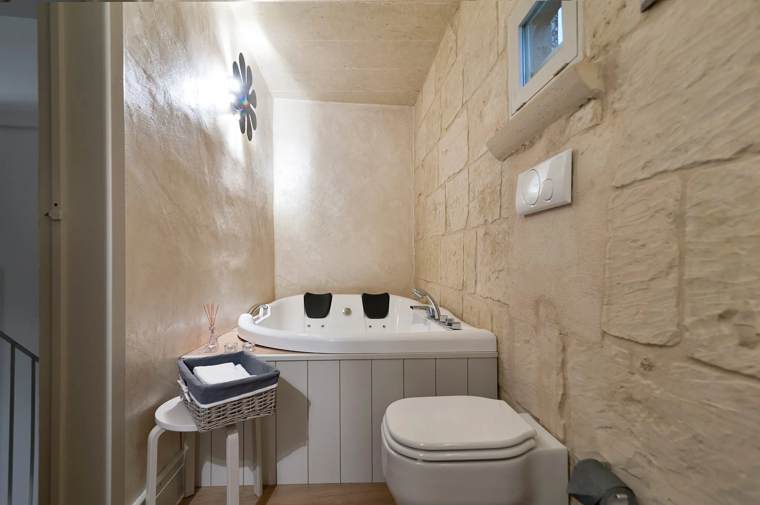 Il bagno della suite Casa Grika: la vasca idromassaggio d’angolo con i poggiatesta scuri, incassata fra le pareti in blocchi di pietra chiara, con l’applique a fiore accesa e una finestrella alta.