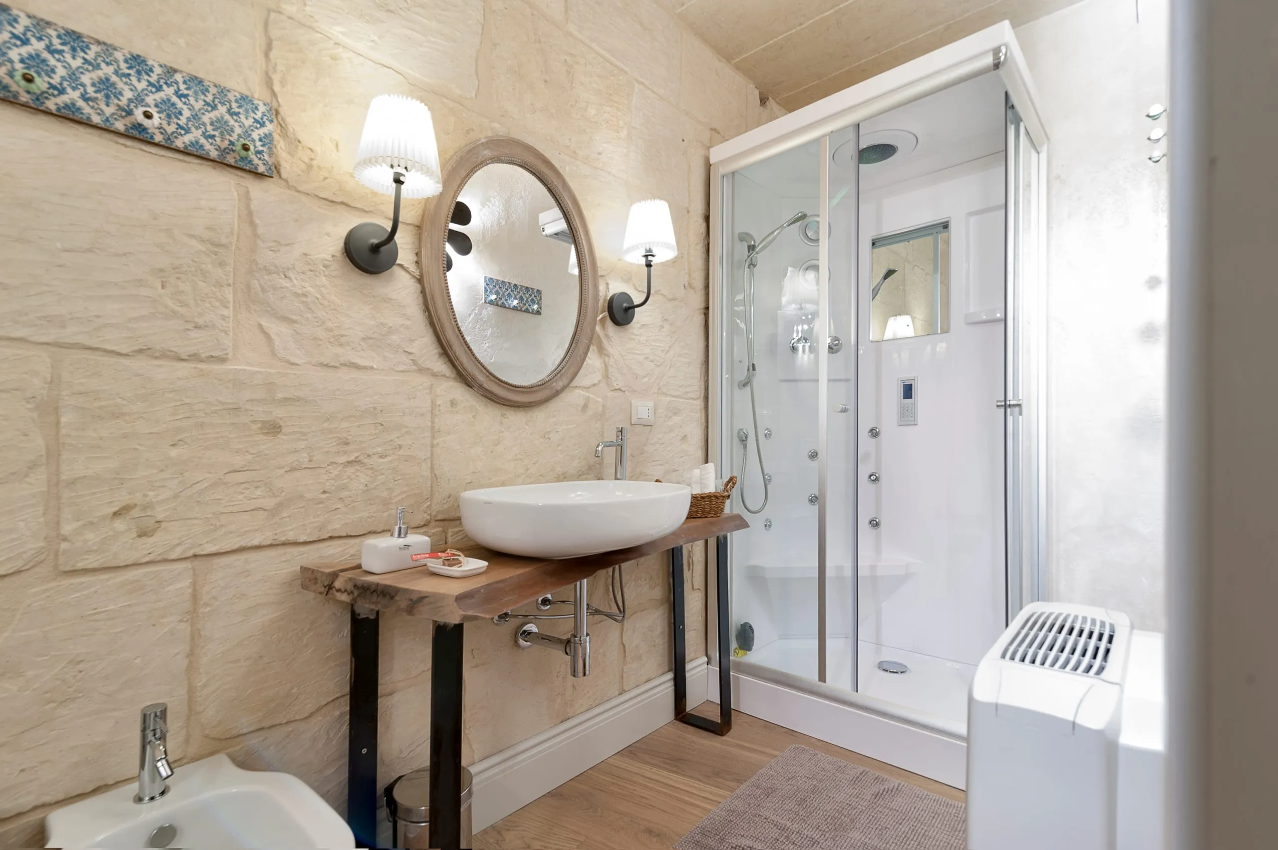 Il bagno di Casa Grika: il lavabo in ceramica su una mensola di legno, lo specchio tondo, la pietra a vista e la cabina doccia.