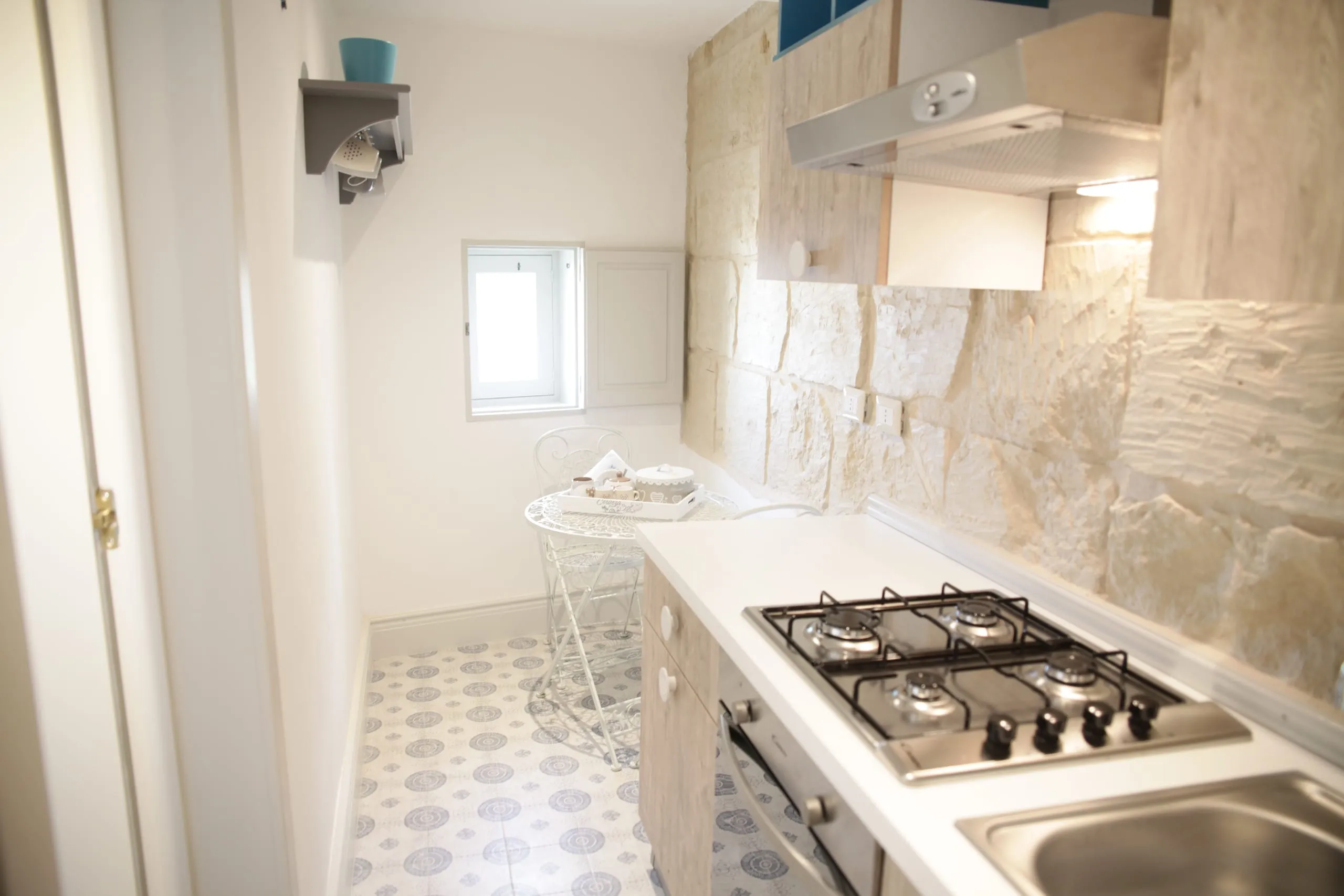 L’angolo cottura di Casa Grika: il piano a gas e il lavello nel muro di pietra, le cementine azzurre e la finestrella in alto.