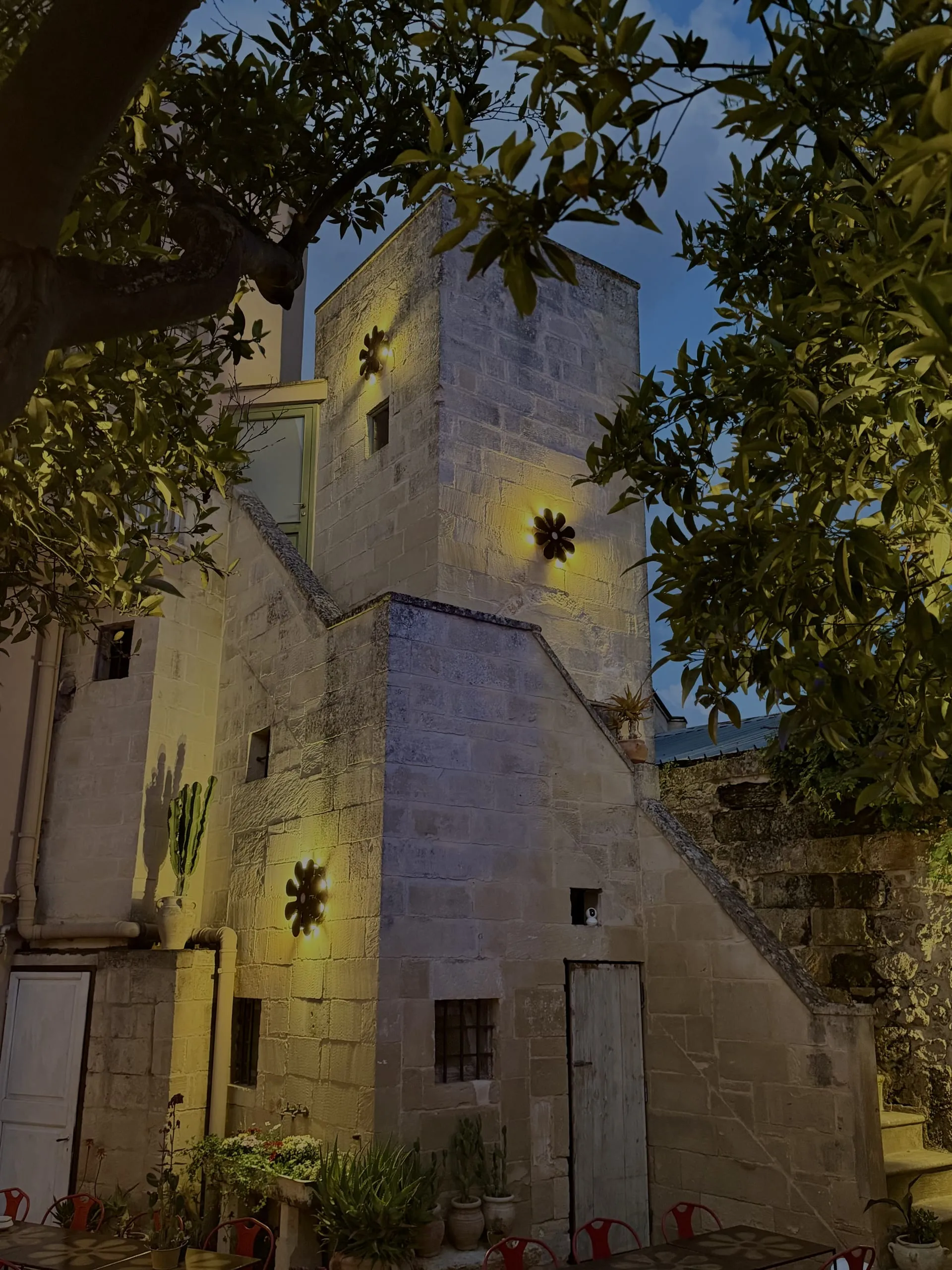 Il cortile interno di Palazzo Mandurino all’imbrunire: le pareti di pietra, le appliques a fiore accese, gli aranci in primo piano e i tavolini con le sedie rosse.