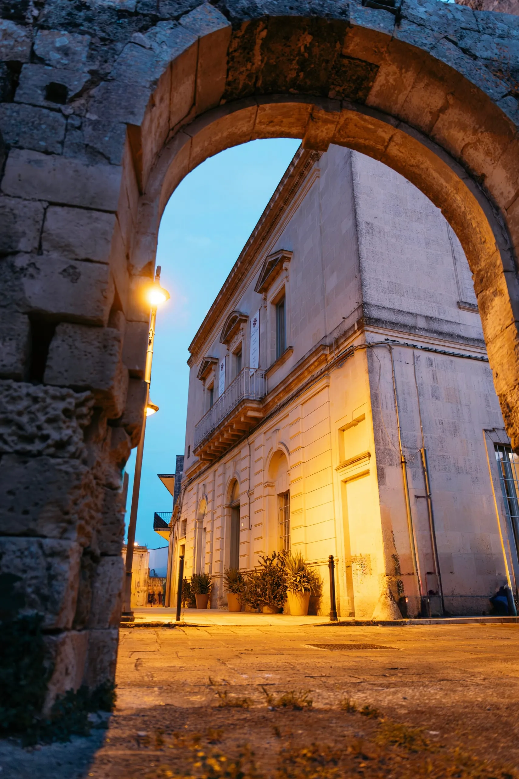 Palazzo Mandurino di sera visto sotto un arco in pietra: il lampione accende la facciata, le piante ai piedi del portone e il cielo ancora blu.
