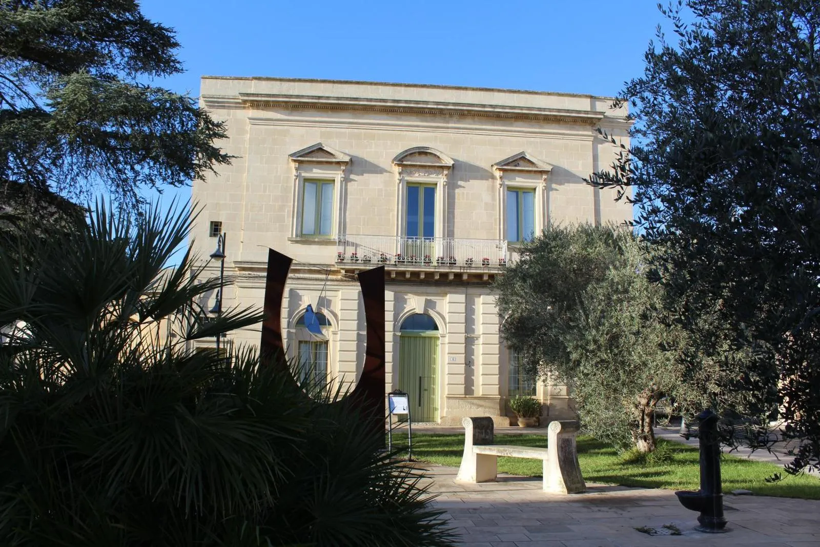 Palazzo Mandurino visto dal giardino della piazza: la facciata in pietra leccese coi tre timpani sulle finestre alte, il balcone in ferro battuto fiorito di gerani e il portale verde ad arco, fra un ulivo e una palma nana, con una scultura in acciaio corten in primo piano.