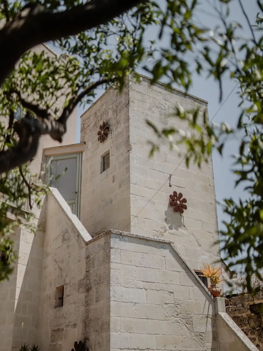 Il volume in pietra di Palazzo Mandurino visto dal basso fra i rami di un ulivo: due fiori in ferro brunito sulla parete chiara, una porta a vetri e la scala esterna.