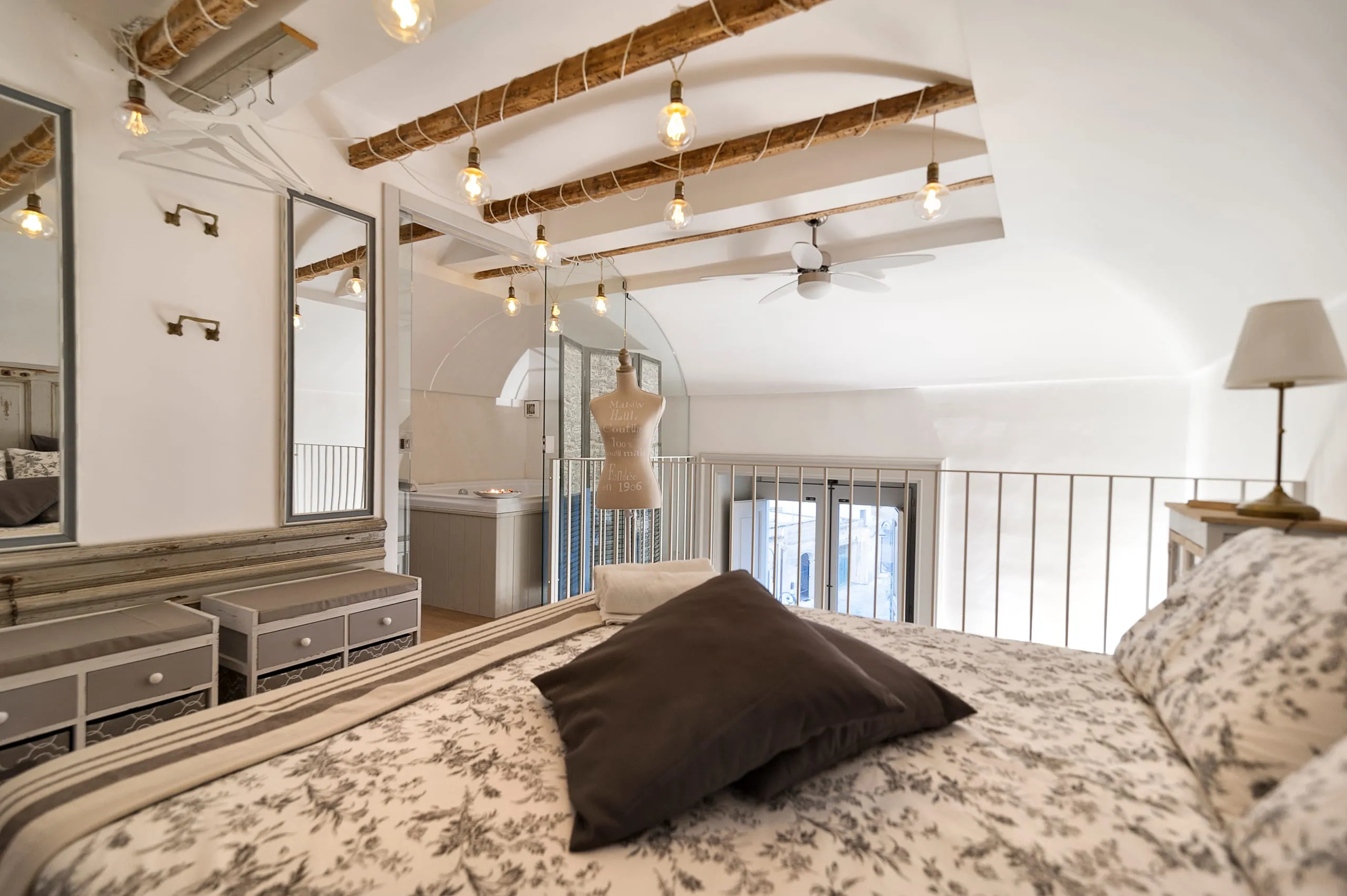 Il soppalco della suite Flora: il letto con la coperta a fiori grigi, un manichino da sartoria accanto alla ringhiera di ferro, le travi di legno e le lampadine appese.
