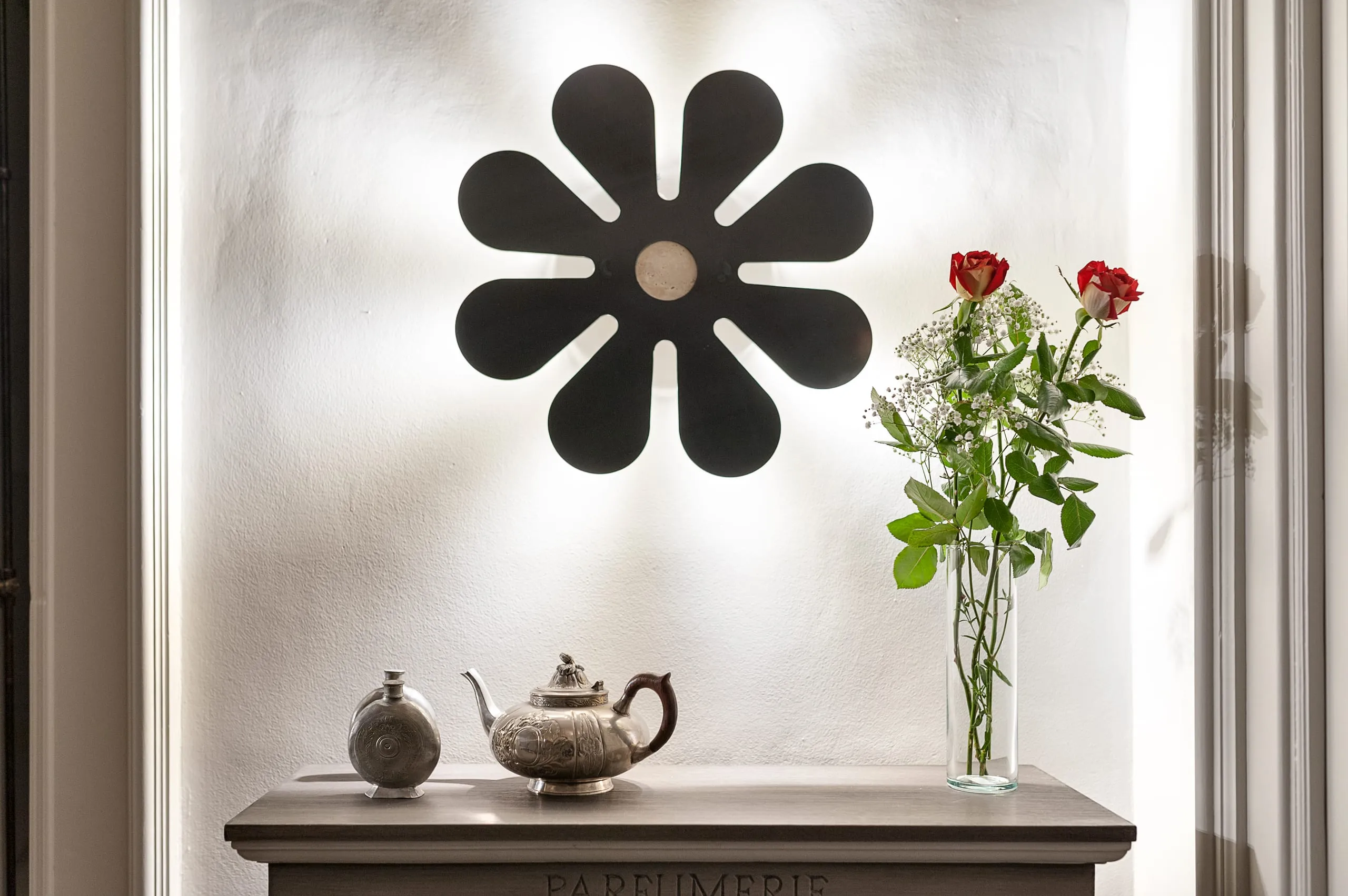 Il fiore nero del marchio alla parete della suite Flora, sopra una console con una teiera di ceramica e un vaso di rose rosse.