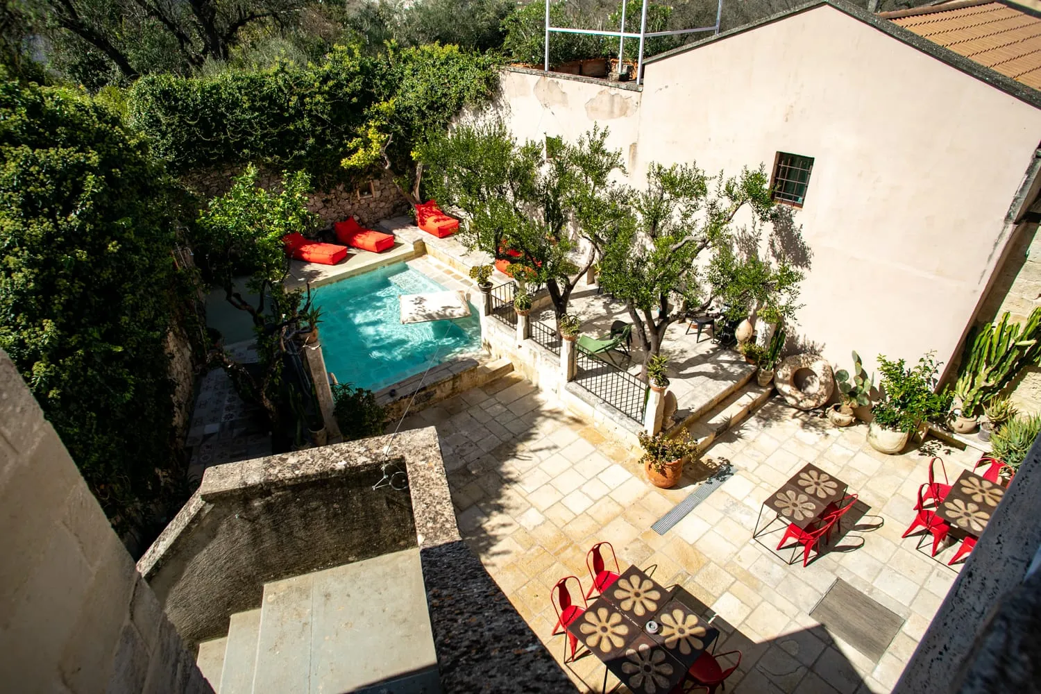 Il giardino di Palazzo Mandurino visto dall'alto: la piscina turchese incassata nella pietra, i cuscini rossi sul bordo, l'ulivo che ombreggia il selciato e i tavolini di ferro con le sedie rosse.