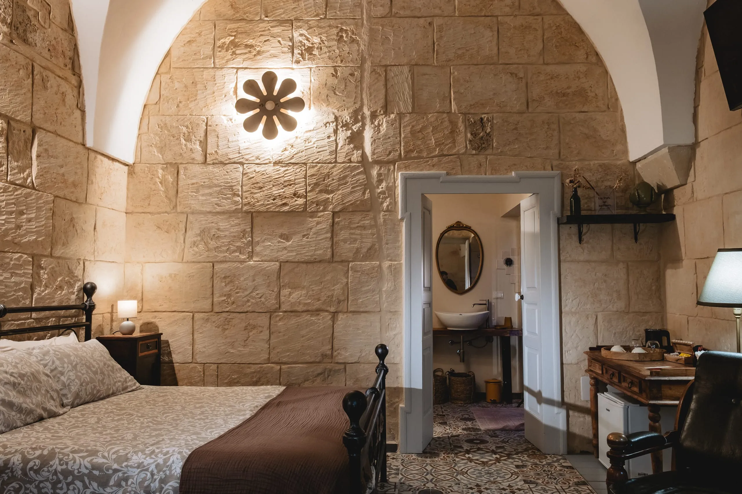 La parete in blocchi di pietra chiara della suite Kali Nifta con l’applique a fiore accesa al centro; a destra la porta aperta sul bagno con lo specchio ovale dorato e il lavabo, e la scrivania di legno con la poltrona di pelle.