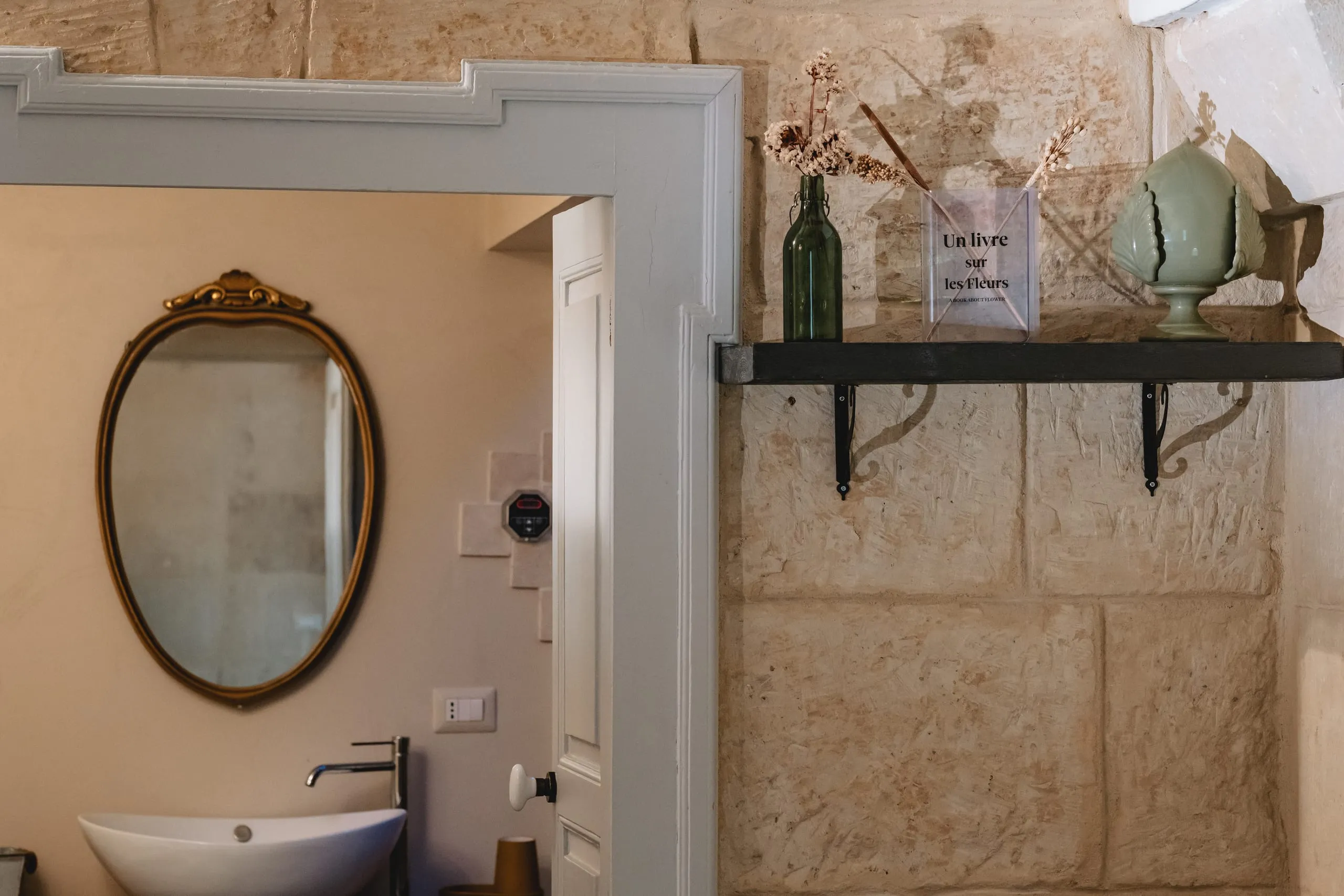 Il bagno di Kali Nifta: lo specchio ovale con la cornice dorata, il lavabo e la mensola scura sul muro di pietra.