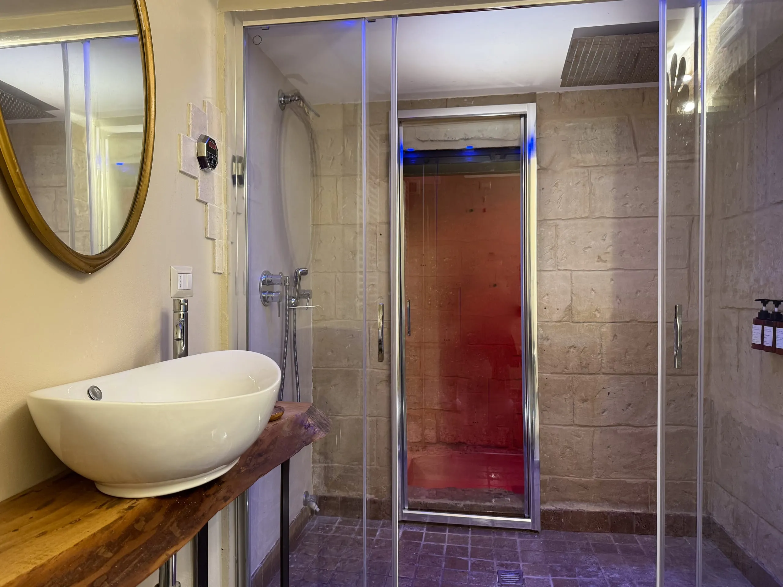 Kali Nifta: il lavabo in ceramica su una mensola di legno e, dietro il vetro, il bagno turco illuminato di rosso.