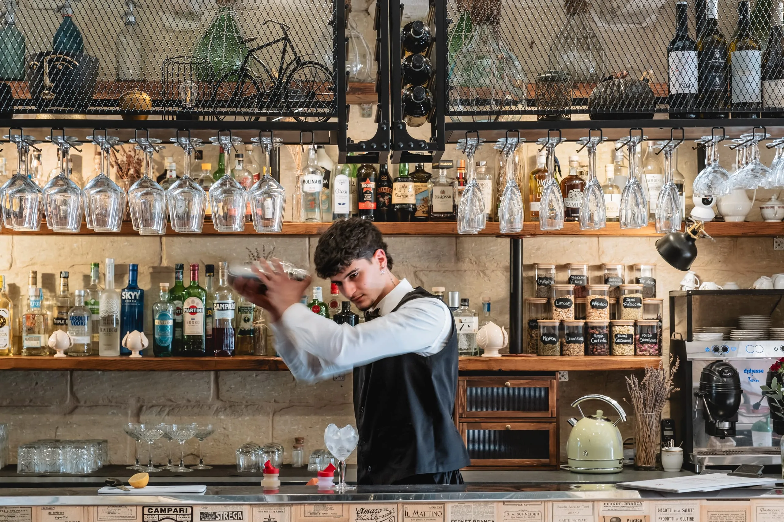 Il bancone del lounge bar: il barman agita lo shaker davanti alle mensole di bottiglie, ai bicchieri appesi e ai barattoli degli infusi.
