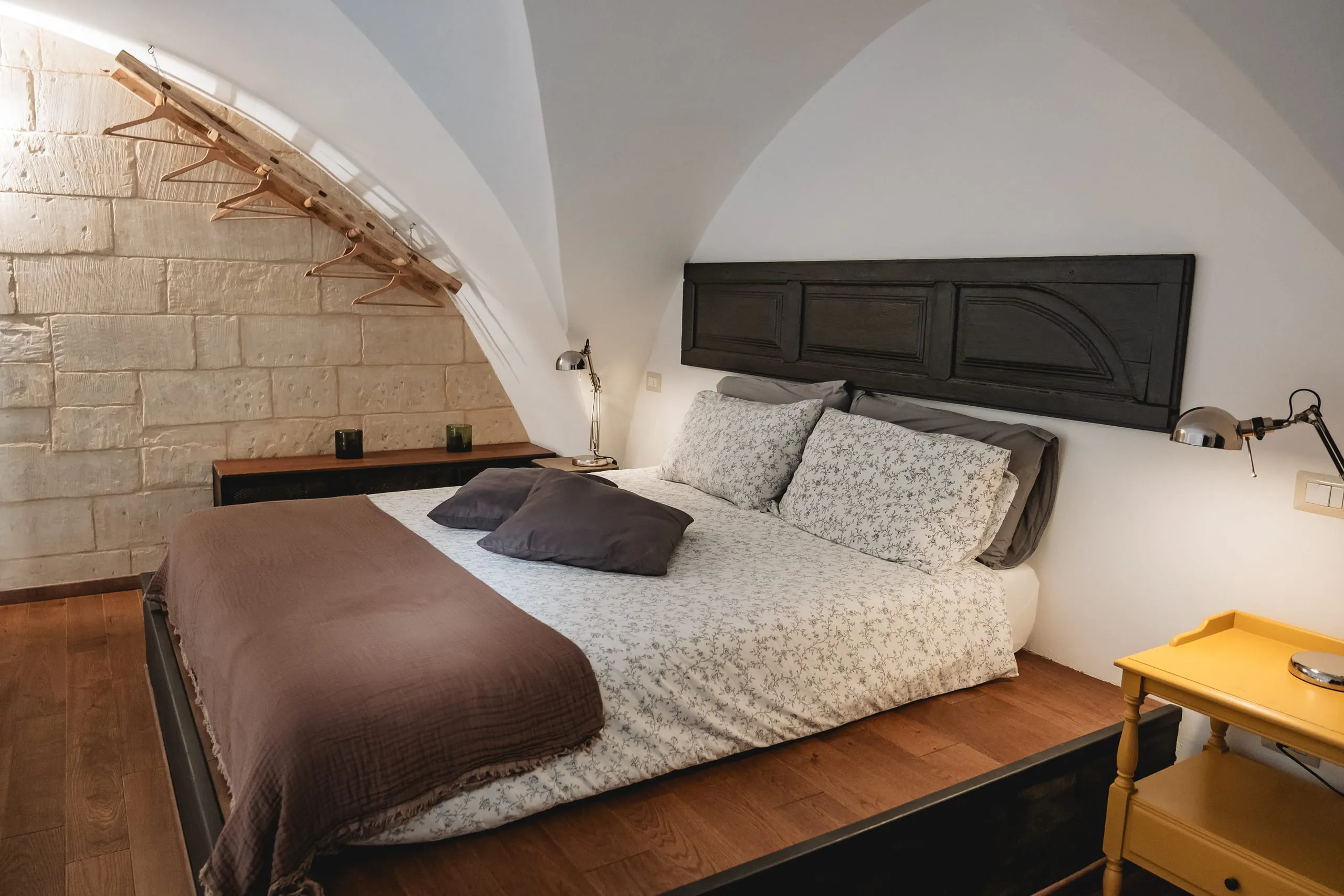 La zona notte della suite Lu Rusciu te lu Mare sotto la volta a crociera bianca: il letto con la testiera ricavata da una vecchia porta scura, il parquet, un comodino giallo e gli appendiabiti di rame alla parete in pietra.