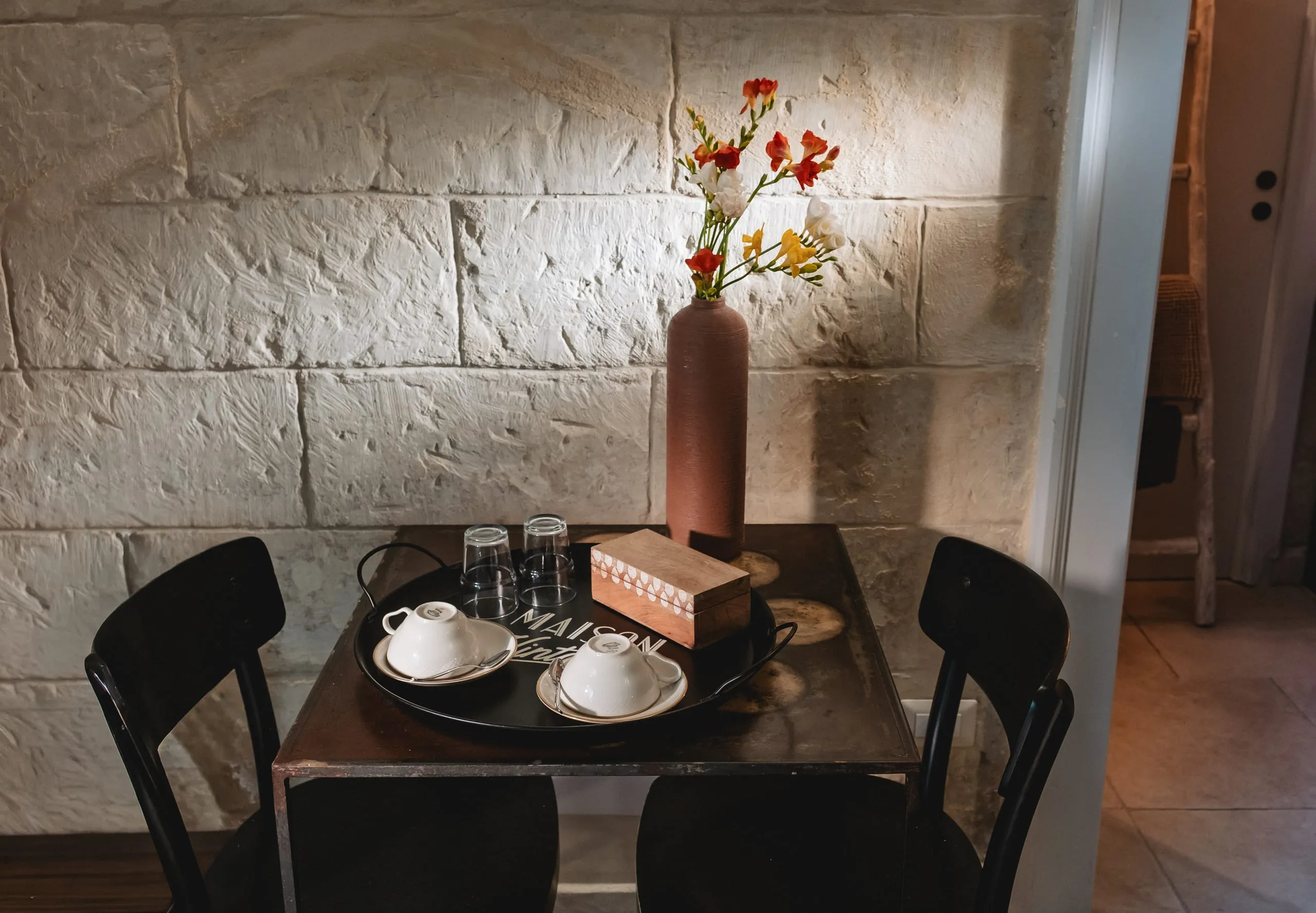 Il tavolino della colazione nella suite Lu Rusciu te lu Mare: due tazze bianche su un vassoio, i bicchieri, un vaso di terracotta con fiori rossi e gialli, contro la parete in pietra chiara.
