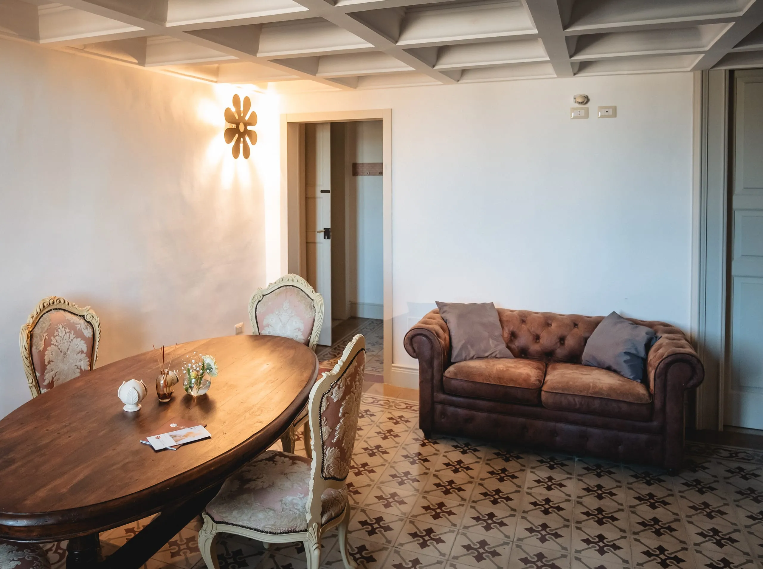 Il salotto della suite Magister: il tavolo ovale di legno scuro con due sedie intagliate, un divano Chesterfield di pelle marrone, il soffitto a cassettoni bianco e il pavimento a cementine.