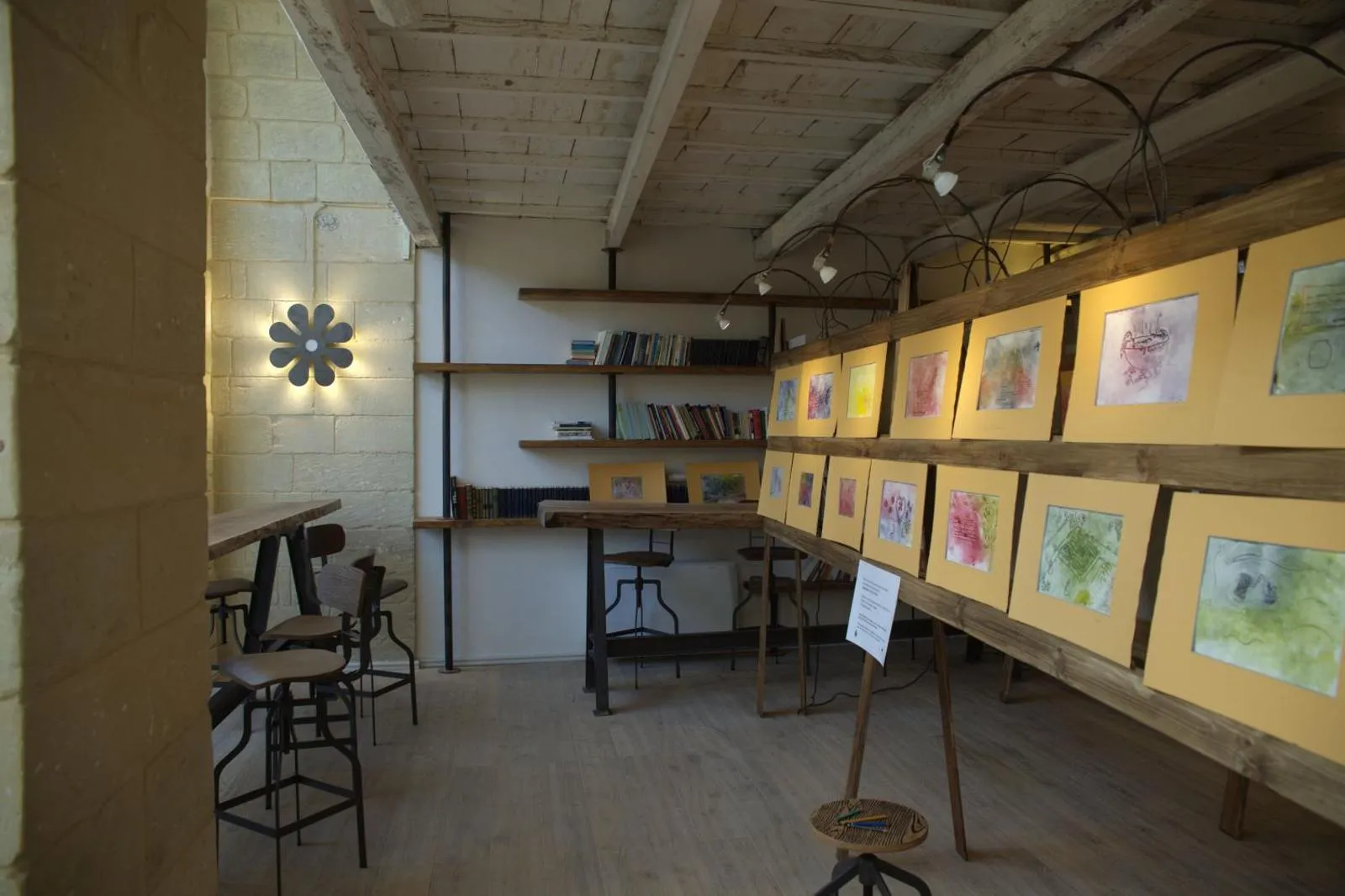 Una mostra allestita nella sala: una fila di acquerelli incorniciati in cartoncino giallo su cavalletti di legno, i faretti sugli archi di ferro battuto, gli scaffali di libri e l’applique a fiore accesa sul muro in pietra.