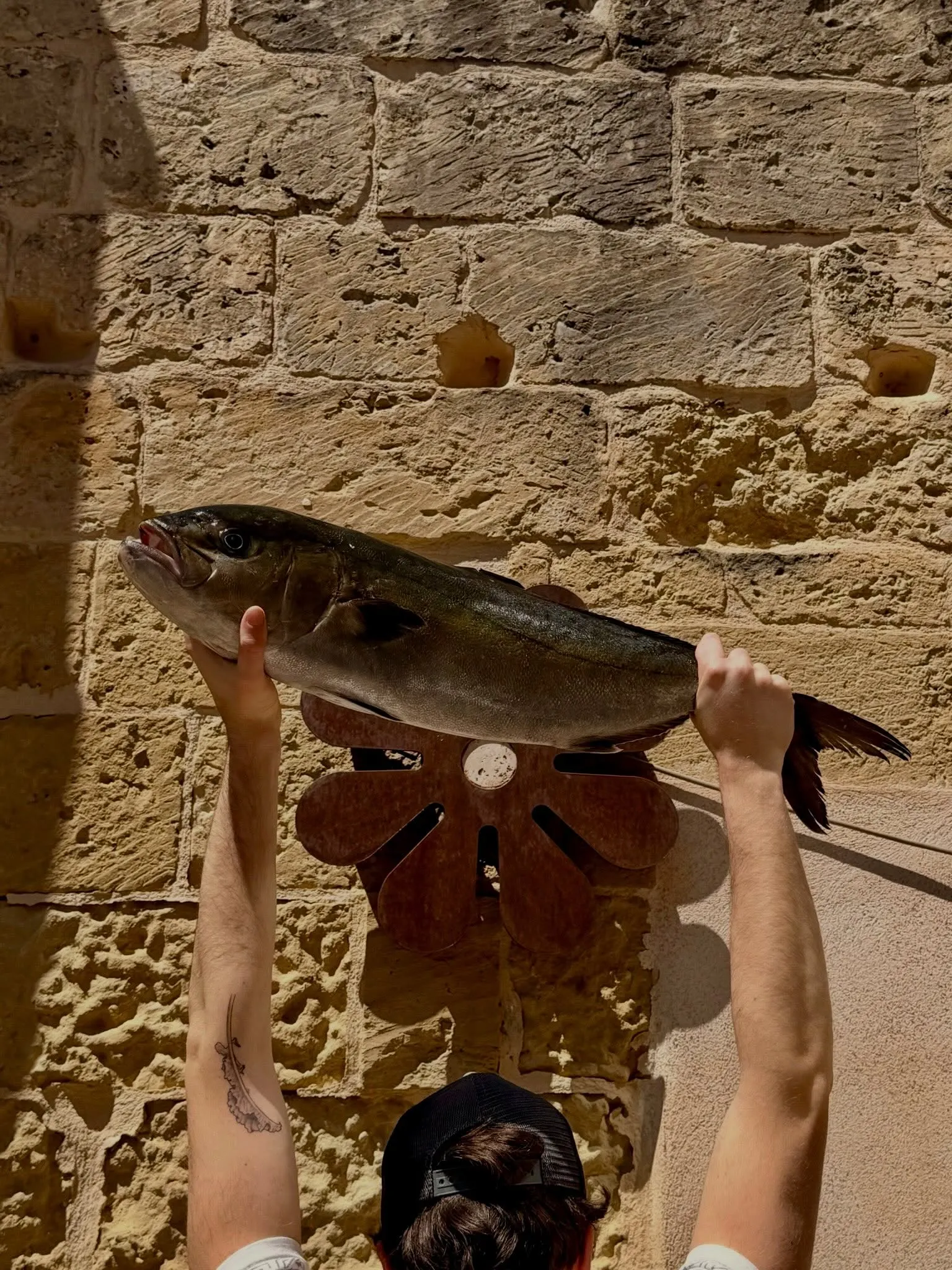 Un cuoco solleva a due mani un pesce appena arrivato contro il muro di pietra leccese, davanti all’applique a forma di fiore in acciaio corten.