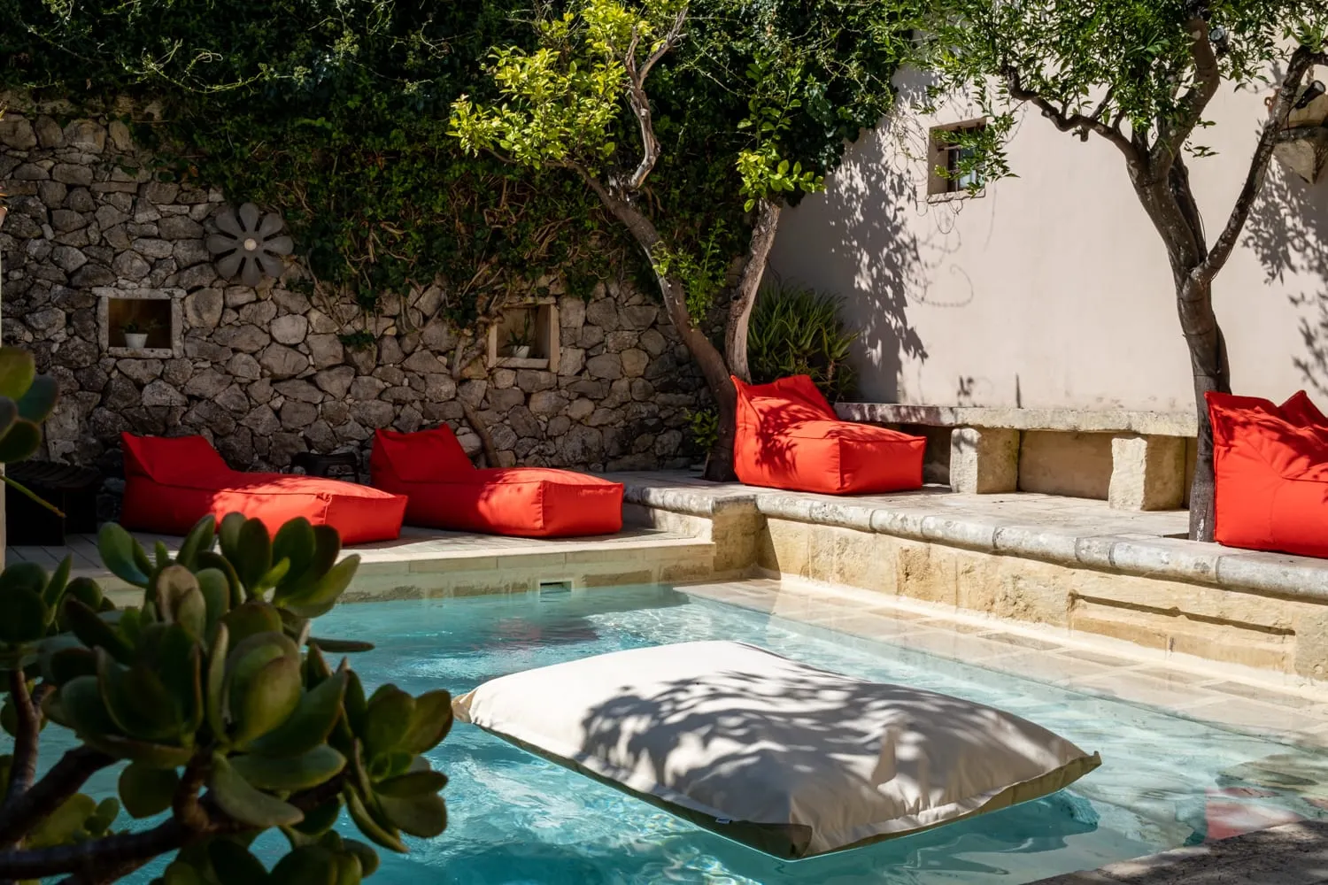 La piscina del giardino in pieno sole: i cuscinoni rossi lungo il bordo in pietra, gli ulivi, il muro a secco e un materassino bianco sull’acqua.