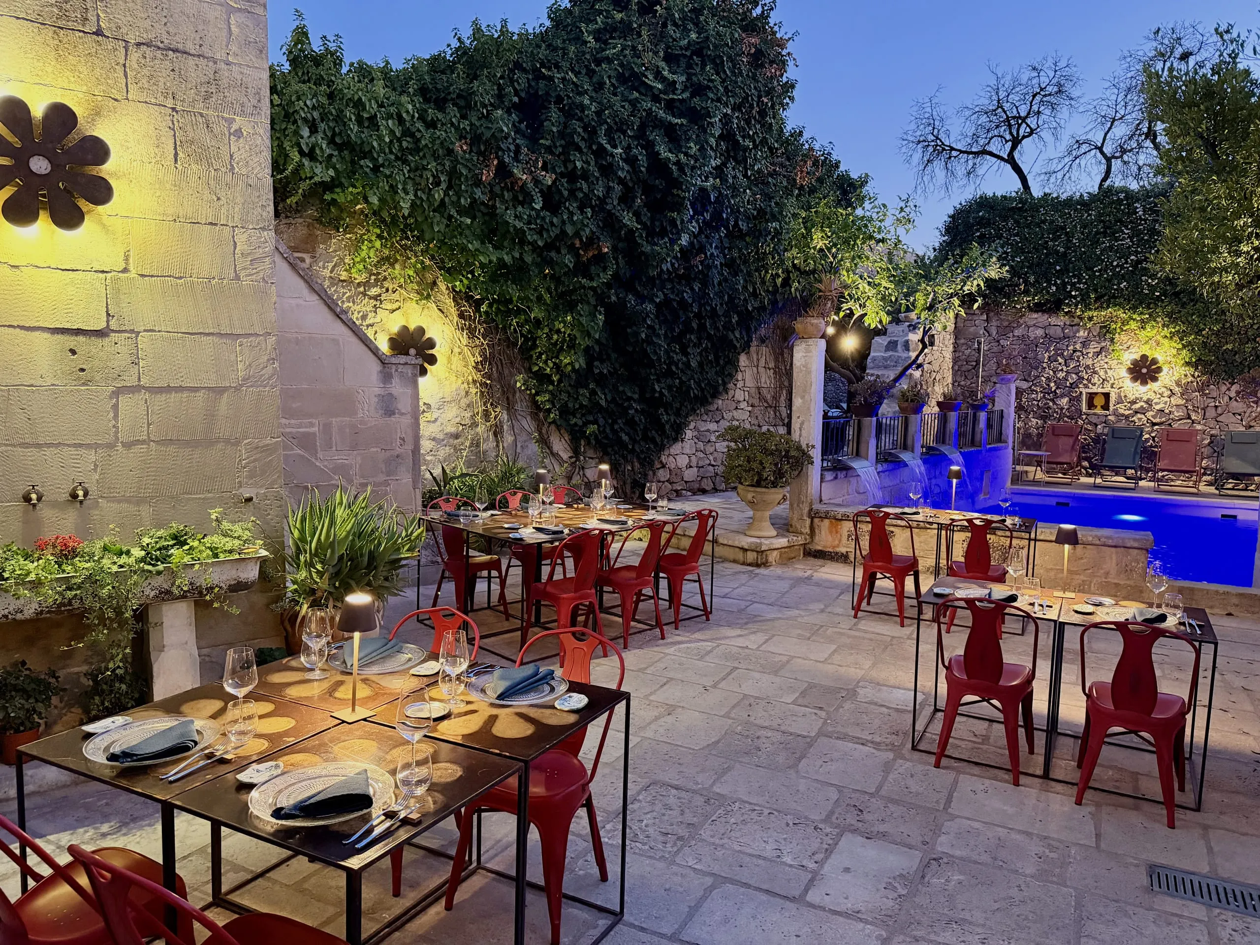 La cena all’aperto nel giardino di sera: le tavole apparecchiate con le sedie rosse, le appliques a fiore accese sul muro di pietra e la piscina illuminata sul fondo.