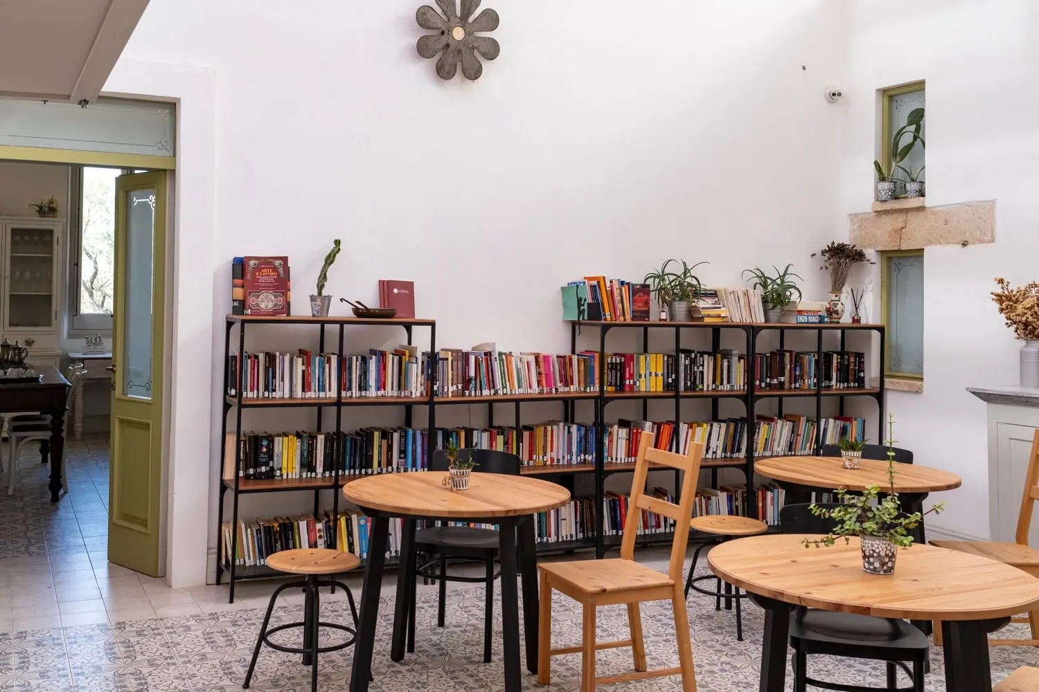La sala della libreria: gli scaffali di libri colorati, i tavoli tondi in legno chiaro, il fiore del marchio sulla parete bianca e la porta verde aperta sulla cucina.