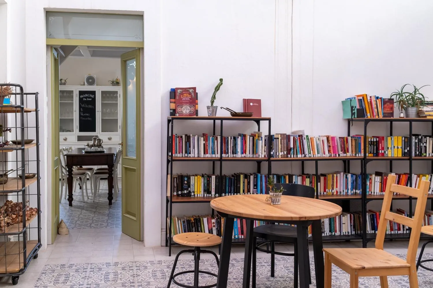 La sala della libreria: gli scaffali di libri colorati, i tavoli tondi in legno chiaro e la porta verde aperta sulla cucina.