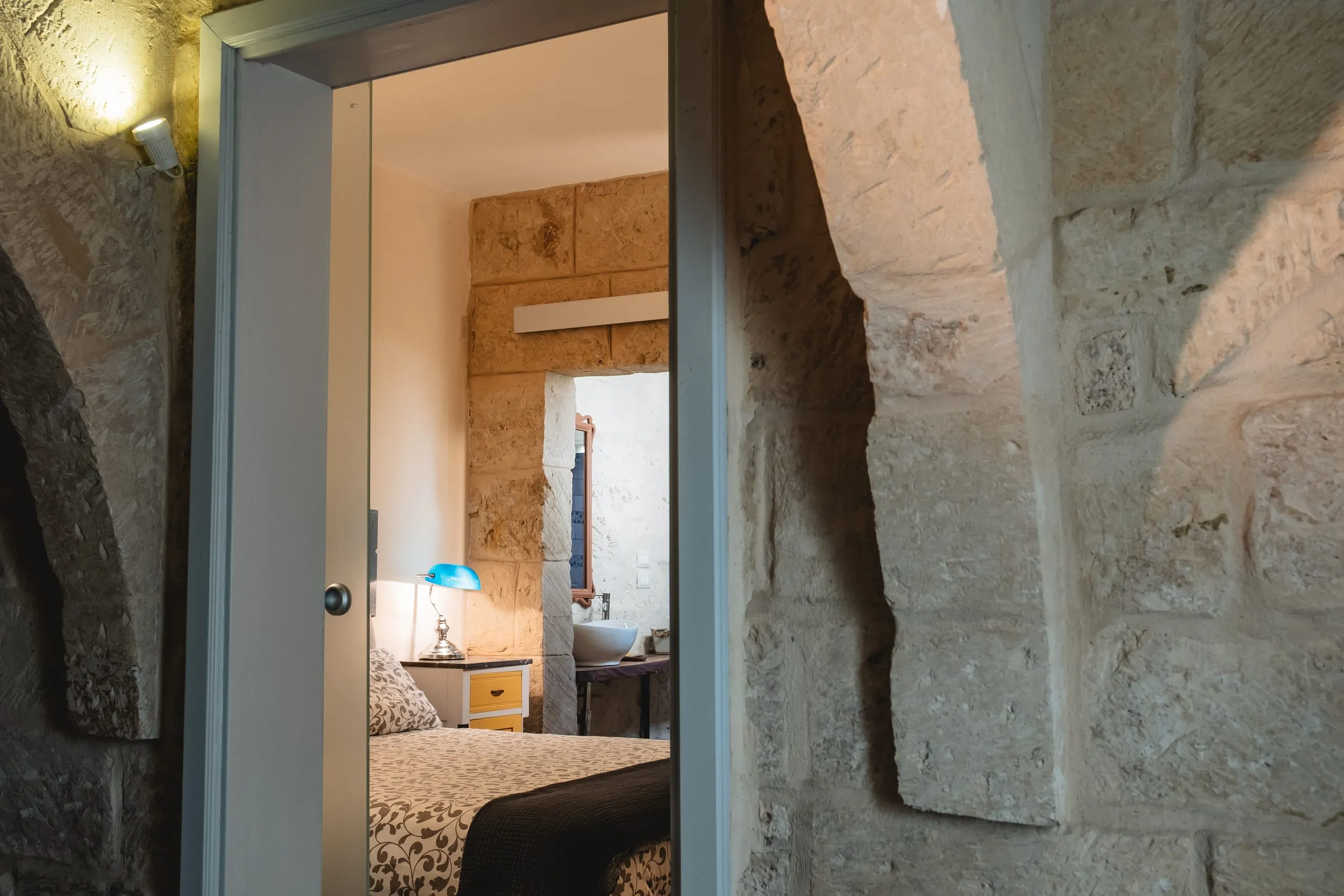 La soglia della suite Sciakuddhi: la porta aperta vista da un ambiente in pietra grezza, che inquadra la camera illuminata con la lampada da tavolo verde smeraldo e il letto oltre la soglia.