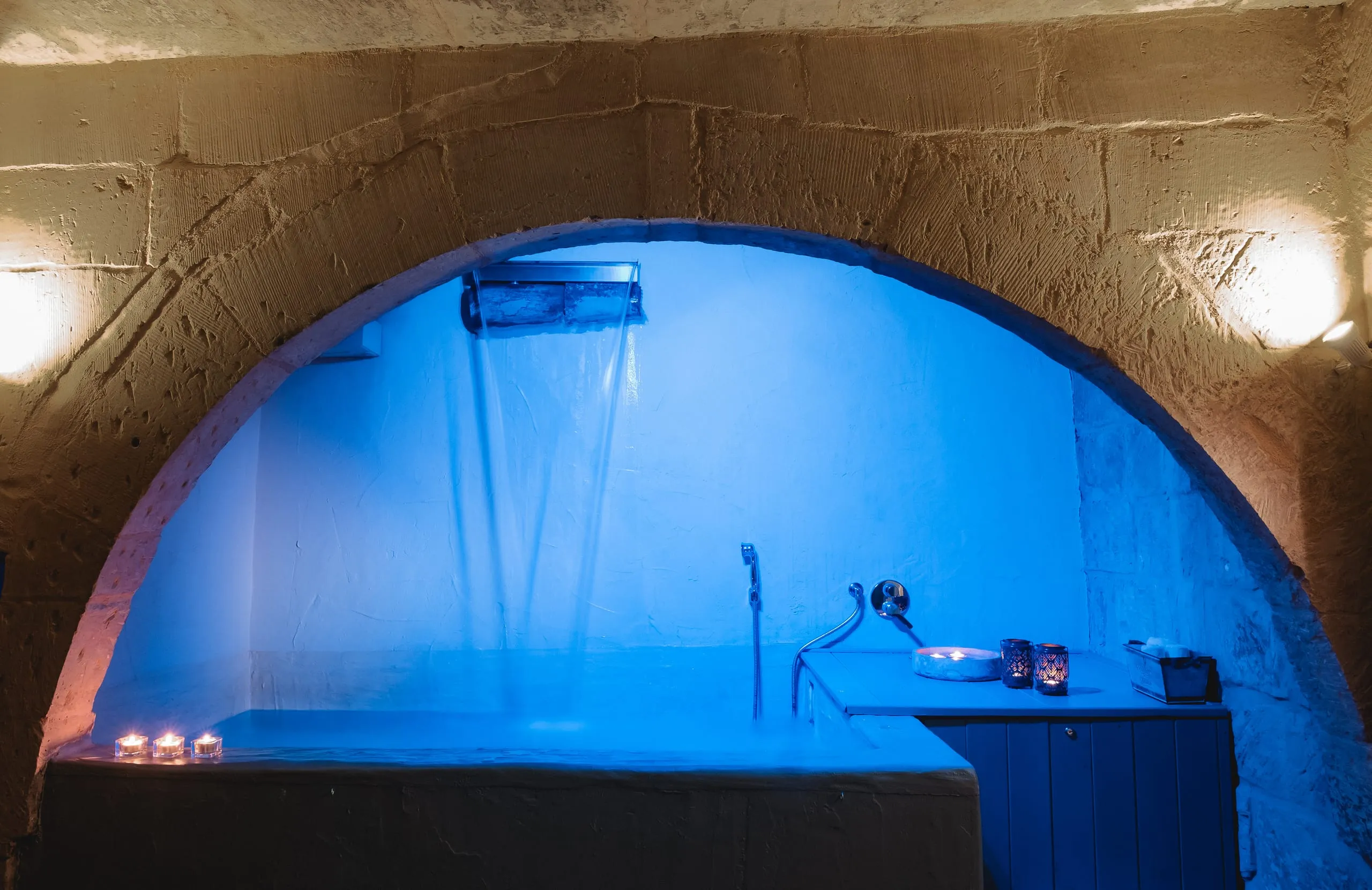 La vasca idromassaggio della suite Sciakuddhi, dentro una nicchia ad arco scavata nella pietra e illuminata di azzurro, con la cascata d’acqua che scende dalla bocchetta a lama e le candele accese sul bordo.