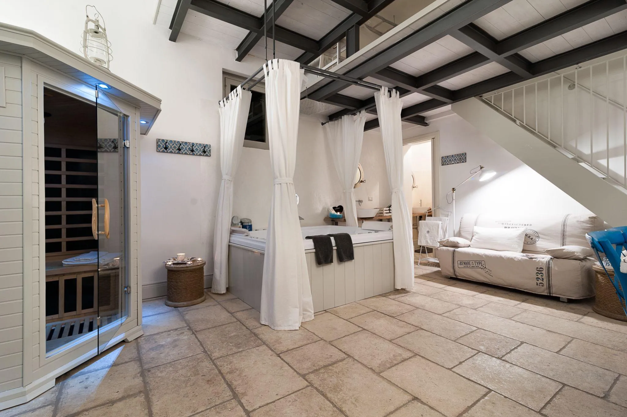 Una suite con la spa privata nel sottotetto: la cabina della sauna accesa, la vasca idromassaggio dietro le tende di lino e il divano sotto le travi.