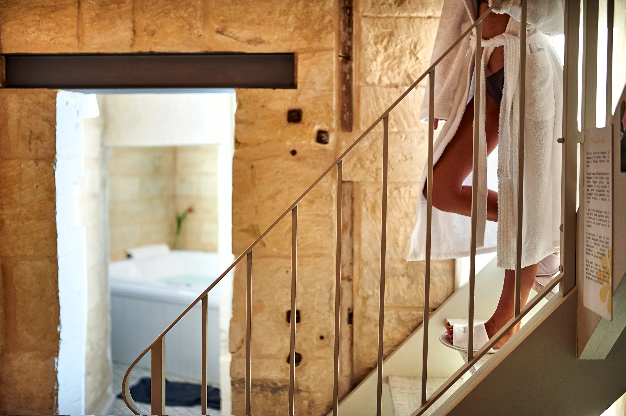 La scala della spa: pietra leccese a vista, la ringhiera sottile e un ospite in accappatoio che sale; oltre la porta, la vasca idromassaggio.