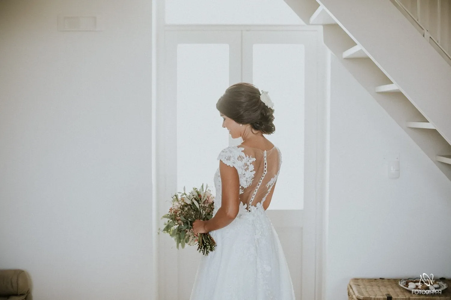 Una sposa di spalle col bouquet, nel suo abito di pizzo, accanto alla scala bianca di una suite.