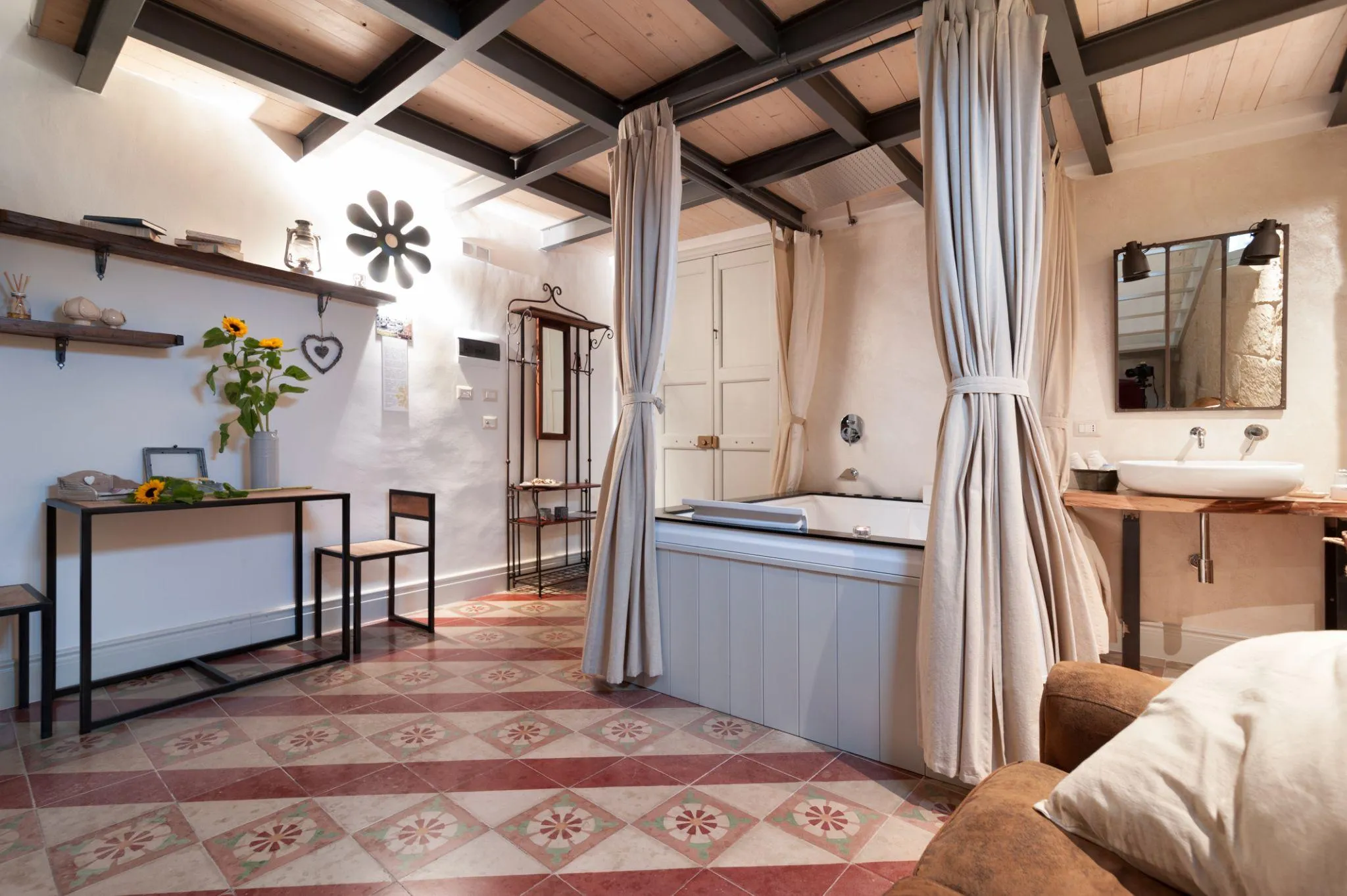 Una suite di Palazzo Mandurino: la vasca dietro le tende di lino, il pavimento a cementine rosse e verdi, i girasoli sulla consolle e l’applique a fiore accesa alla parete.