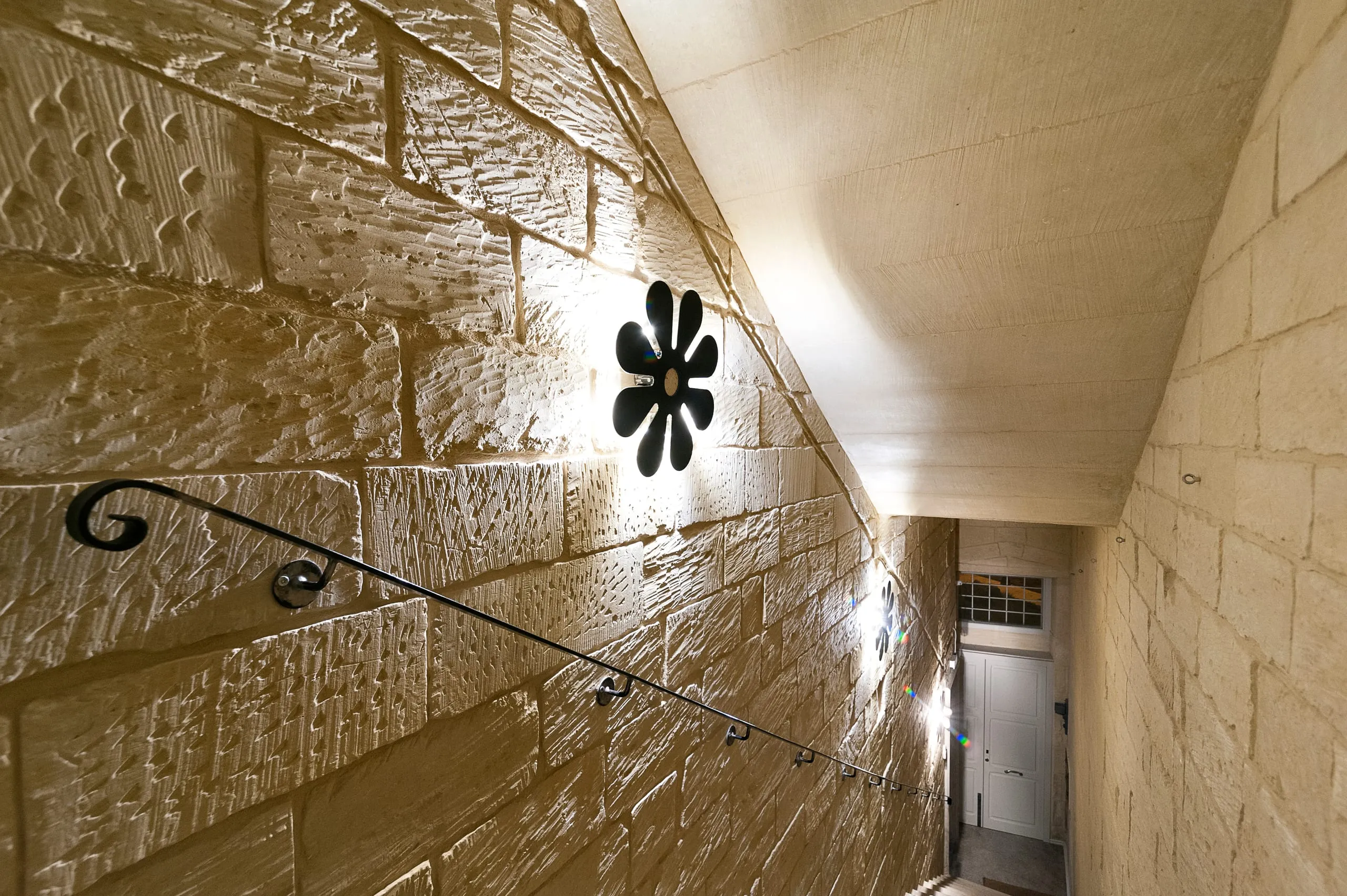 Il vano scala di Palazzo Mandurino: i gradini fra due muri in blocchi di pietra chiara illuminati dal basso, il corrimano di ferro battuto e l'applique a fiore nero accesa sulla parete.