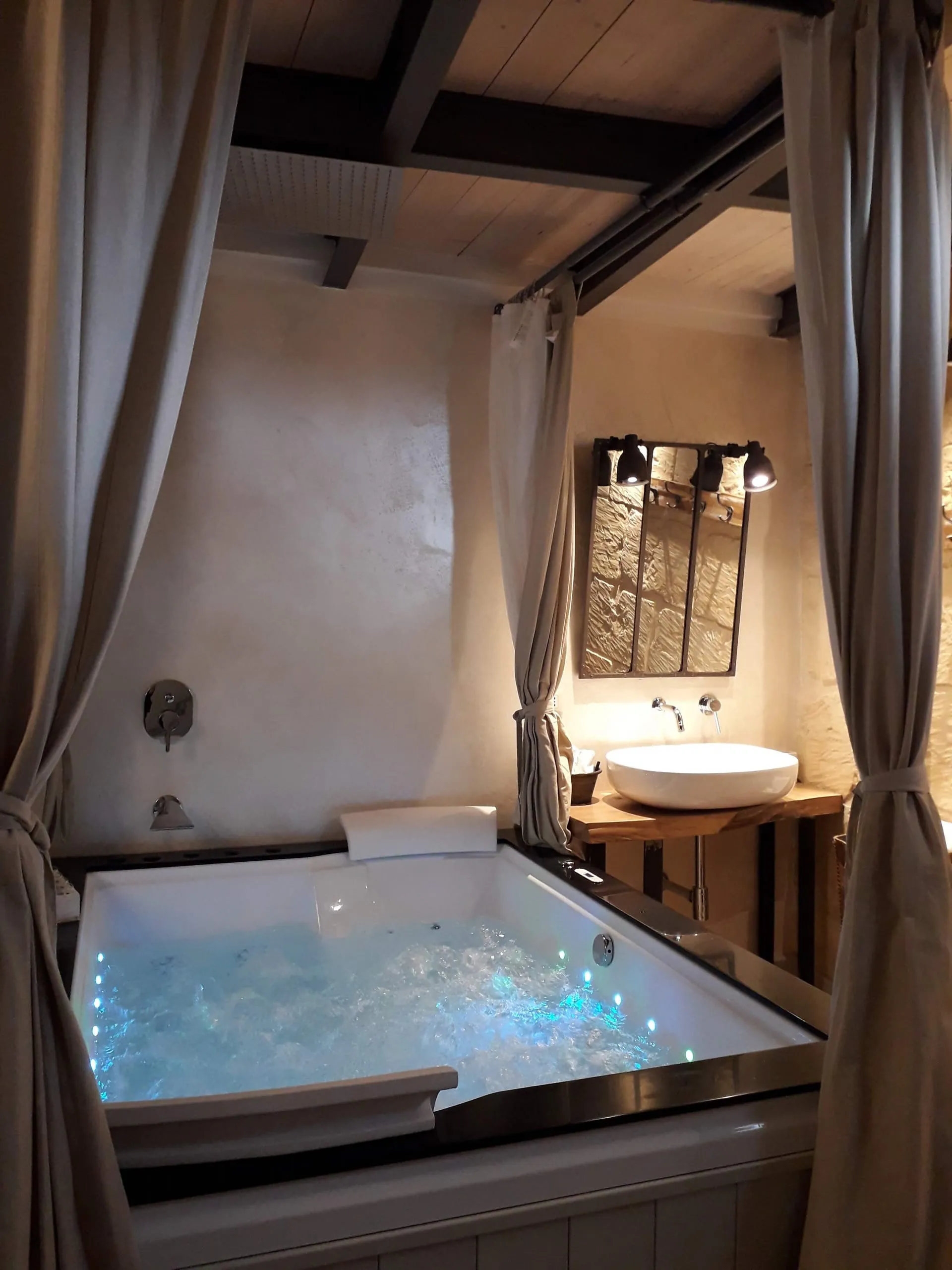 La vasca idromassaggio illuminata fra le tende di lino, sotto le travi, con il lavabo in ceramica e lo specchio a finestra.