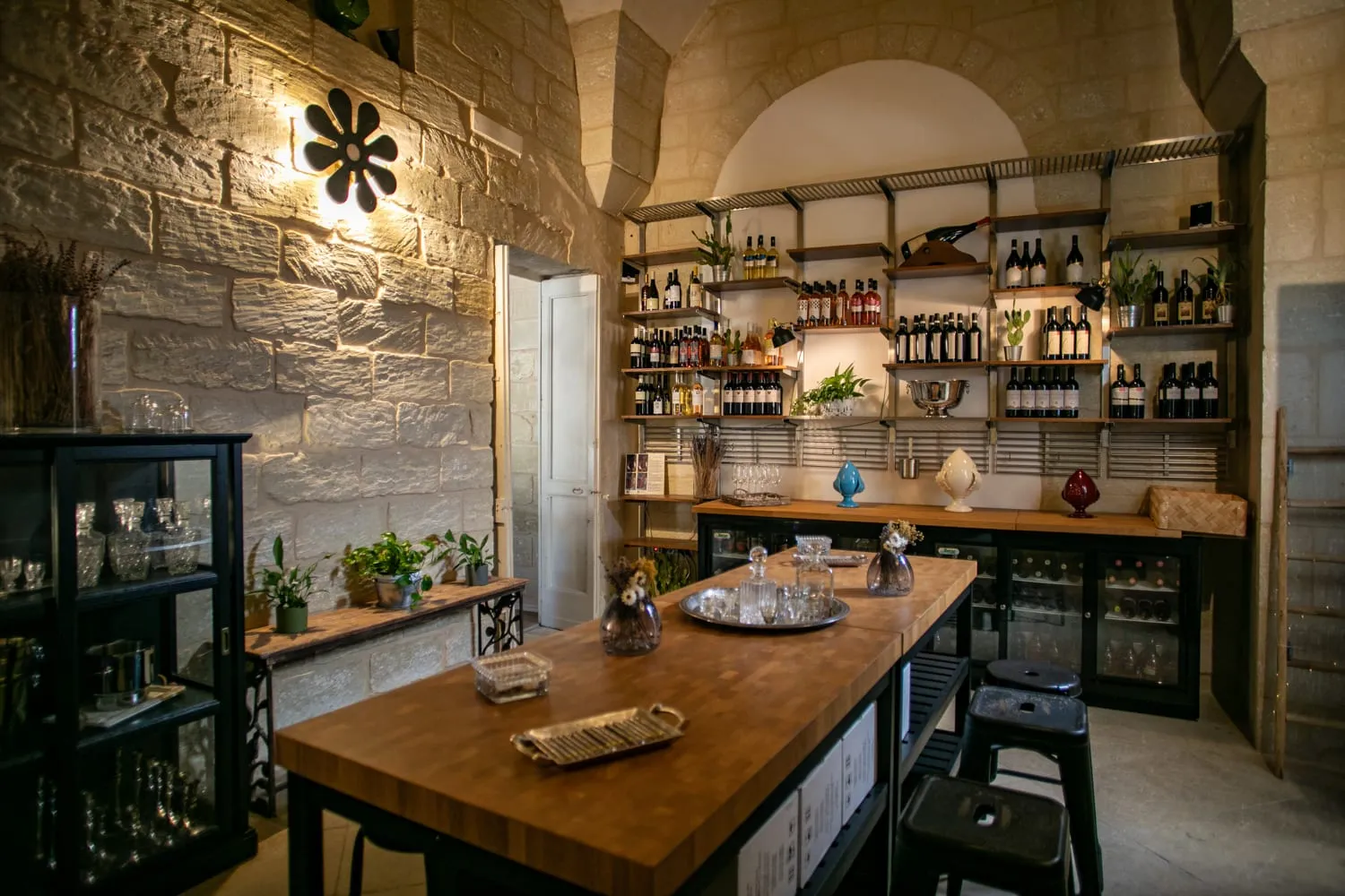 La vineria del Palazzo sotto la volta in pietra: il lungo tavolo in legno, le bottiglie sugli scaffali, i pumi di ceramica e l’applique a fiore accesa sul muro.