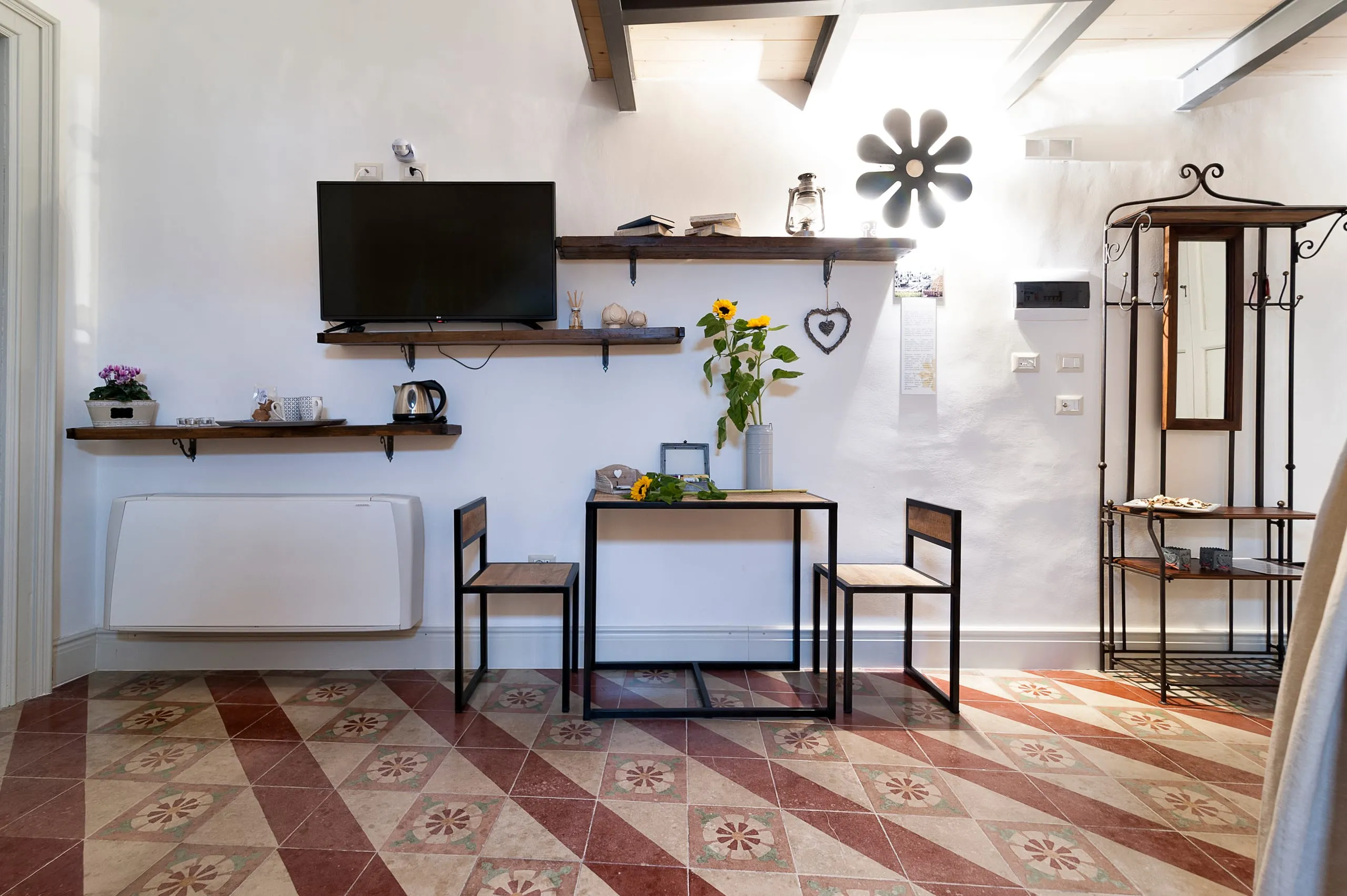 Il salotto della suite Xanti Yaca: il pavimento a cementine rosse e bianche posate in diagonale, un tavolino con due sedie di ferro, l’applique a fiore alla parete e il soffitto a travi di legno.