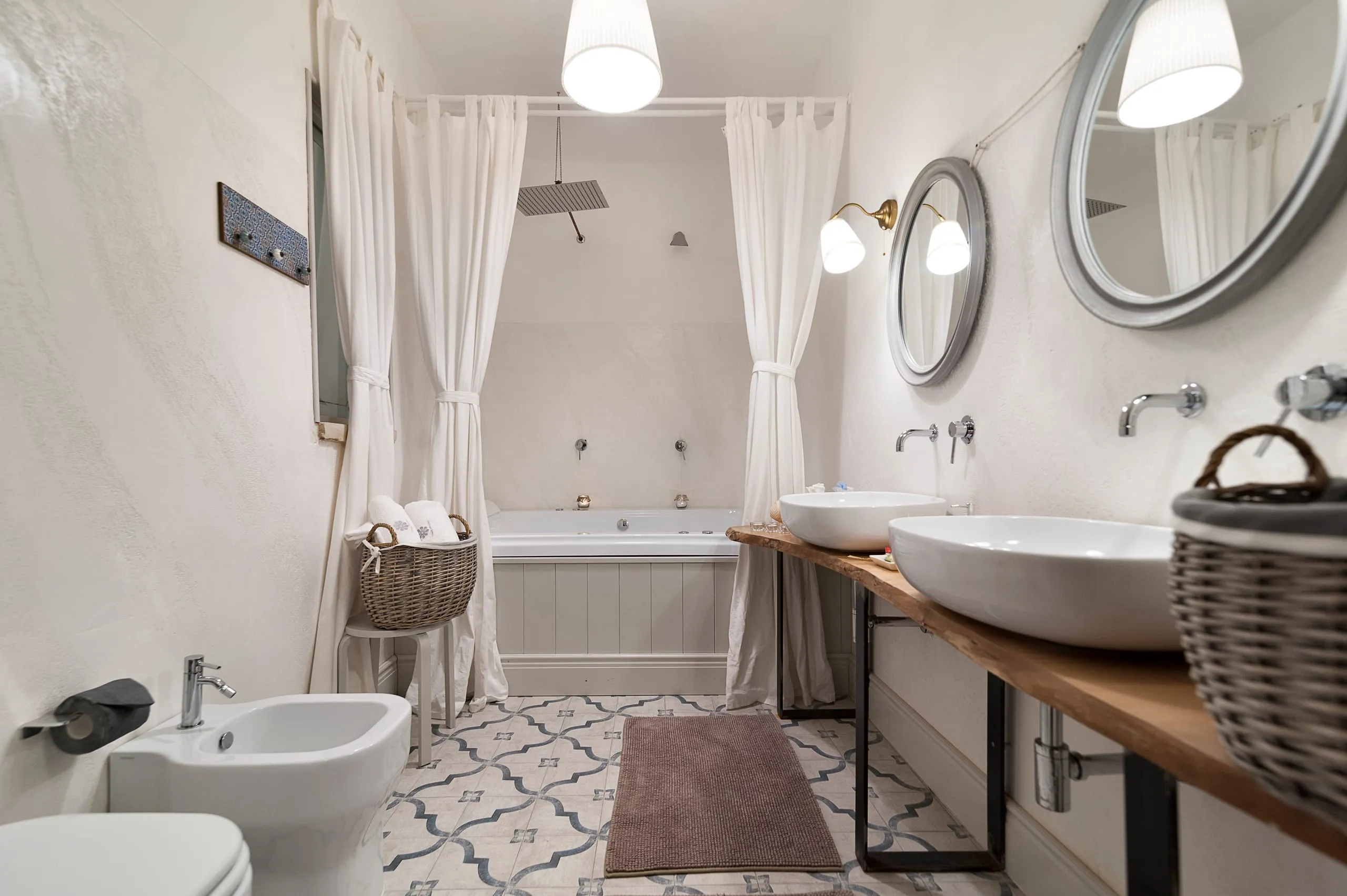 Il bagno della suite Zagareddha: la vasca idromassaggio dietro tende bianche con la doccia a pioggia sopra, due specchi tondi e il doppio lavabo su una mensola di legno grezzo.